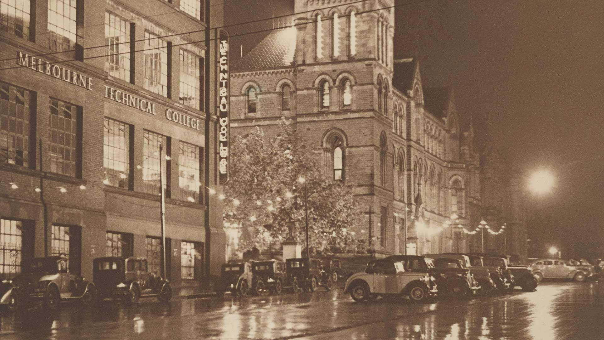 Historical, sepia tone image, of Melbourne Technical College building, at night, with cars parked in front of the building
