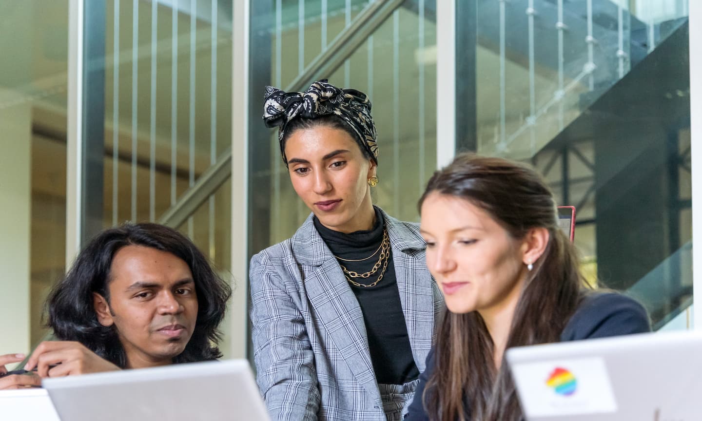 Three people around a laptop wearing professional attire, two sitting, one standing behind.