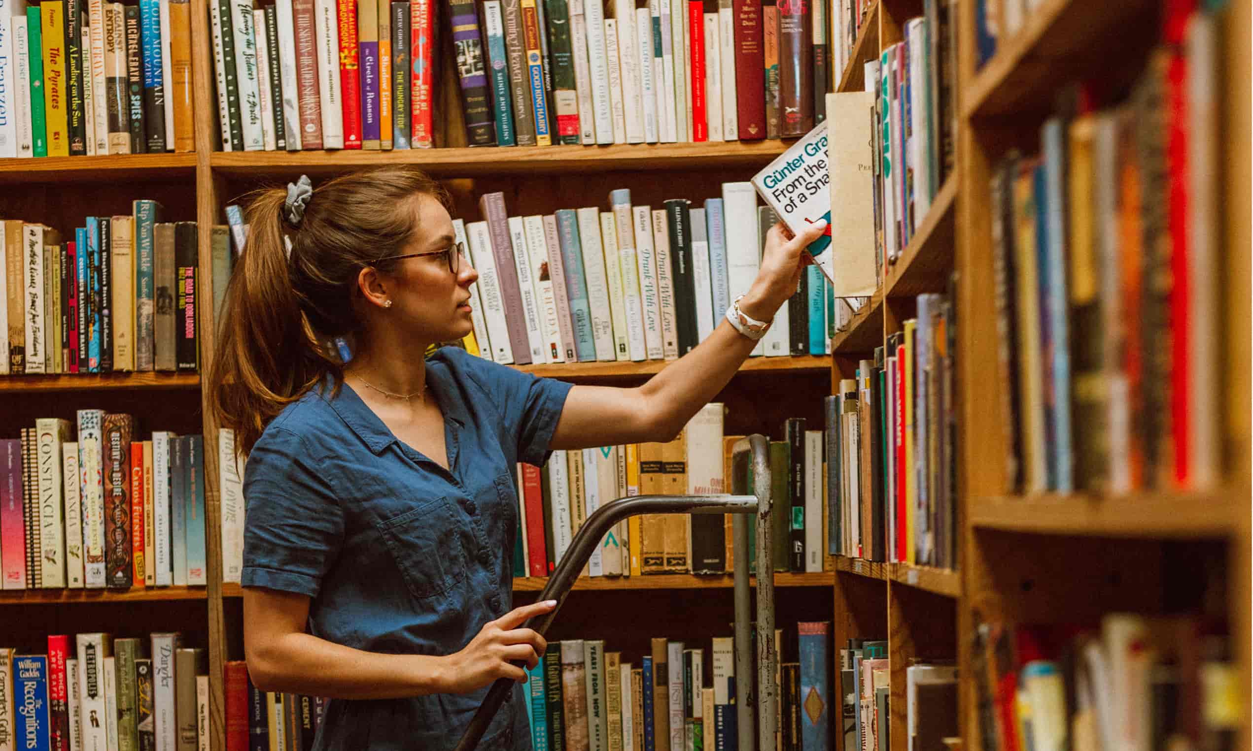 A woman surrounded by shelves filled with books is taking a book off one of the shelves